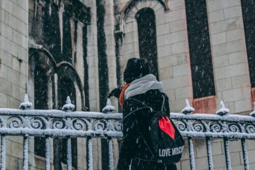 A woman with a scarf gazes at a landmark during snowfall in Paris.